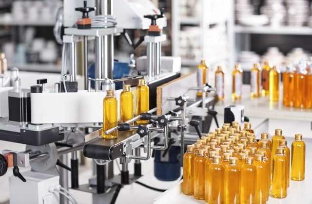 Horizontal shot of cosmetics or pharmacy plant with automated equipment. Transparent plastic bottles filled with yellow substance standing on desk and conveyor line, ready for transportation Horizontal shot of cosmetics or pharmacy plant with automated equipment. Transparent plastic bottles filled with yellow substance standing on desk and conveyor line, ready for transportation